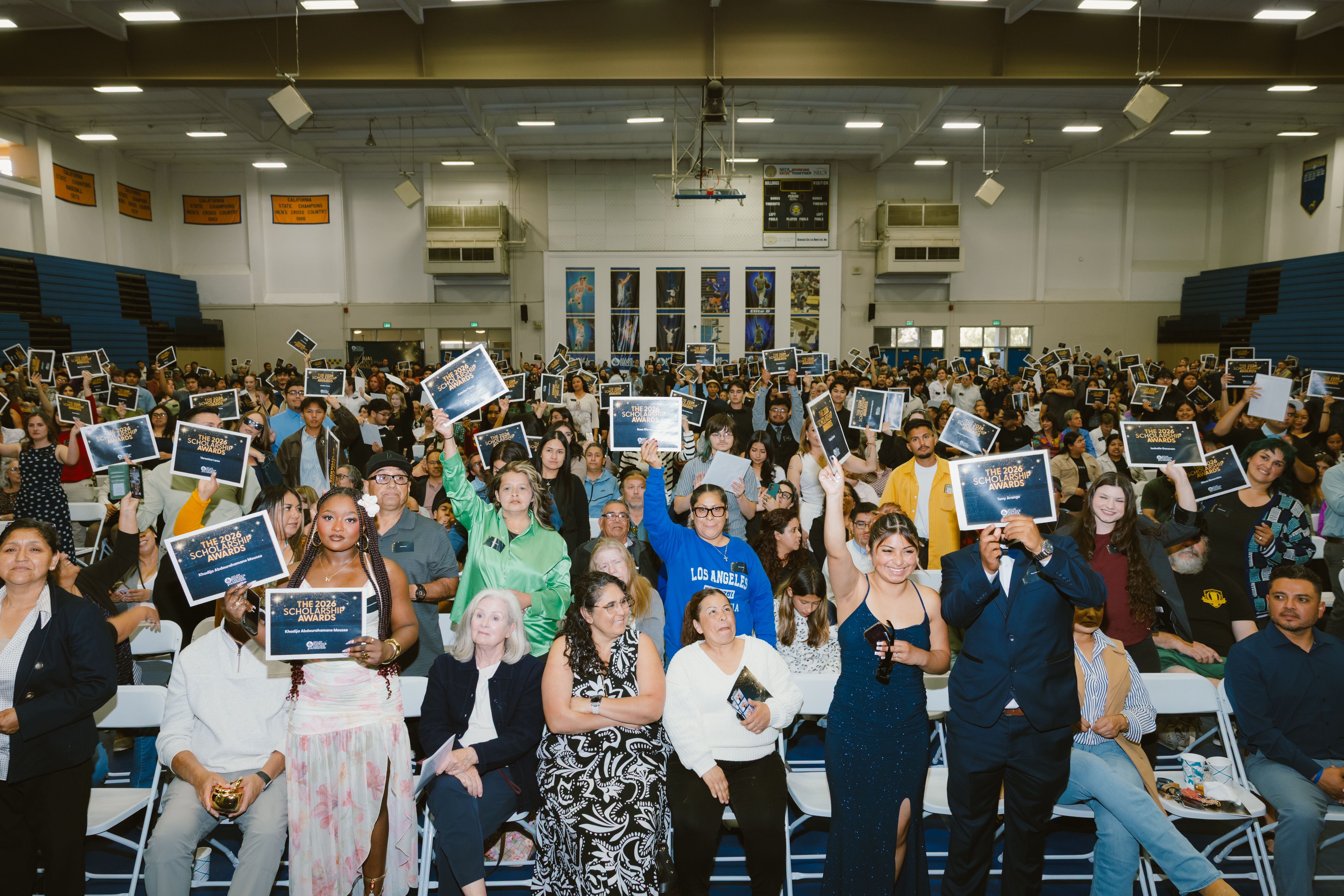Students and supporters gather inside an Allan Hancock College gymnasium during the annual Scholarship Awards ceremony. Many attendees hold up signs recognizing 2026 scholarship recipients while seated families and guests celebrate throughout the packed venue.